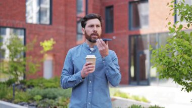 Akıllı telefon kullanan yakışıklı bir iş adamı dışarıda ofis binasının yanında kahve içiyor. Cep telefonu kullanan günlük gömlekli profesyonel bir adam..