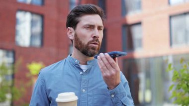 Açık havada akıllı telefon kullanan yakışıklı bir iş adamı. İş, teknoloji, kahve molası. Mavi tişört giyen yetişkin bir uzman, ciddi hissediyor. Şehir arkaplanı.