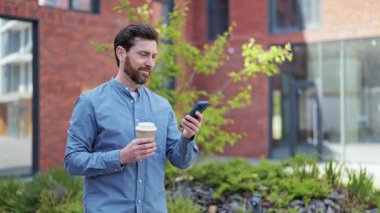 Günlük giysiler içindeki mutlu iş adamı kahve içer ve modern ofisin yakınında akıllı telefon kullanır. Sokakta kahve ve telefonla gezen neşeli bir iş adamı..