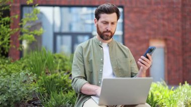 Başarılı bir iş adamı açık havada günlük kıyafetlerle çalışıyor. Bilgisayarı ve telefonu kullanırken kendini mutlu hissediyor ve başarıyı kutluyor..