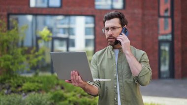 Başarılı iş adamı, akıllı telefonla konuşurken ve dizüstü bilgisayarını tutarken dışarıda kötü haberler verirken hayal kırıklığına uğramış hissediyor. Sıradan kıyafetler giymiş gözlüklü profesyonel bir erkek..