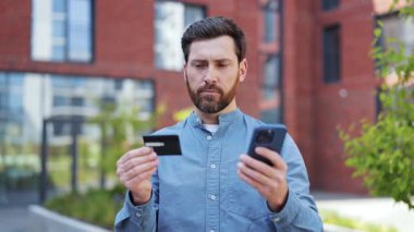 Günlük giysiler içinde yakışıklı bir işadamı akıllı telefon ve kredi kartıyla online ödeme yapıyor. Başarılı adam dışarıdan satın almaktan memnun. Fintech, alışveriş..