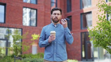 Odaklanmış işadamı kahve içiyor, hoparlörden konuşuyor, ofis binasının yakınında neşe ve heyecan yaşıyor. Sıradan stil.
