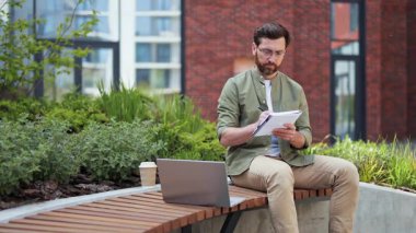 Üretken erkek öğretmen işadamı, günlük kıyafetlerle uzaktan çalışıyor. Dizüstü bilgisayarı olan başarılı bir adam not alır ve çevrimiçi toplantıda konuşur. İş, serbest çalışma, uzaktan çalışma.