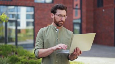 Açık havada dizüstü bilgisayarda çalışırken günlük kıyafetler ve gözlüklerle gülümseyen iş adamının portresi. Modern bina. Başarılı bir iş. Keyfin yerinde. Profesyonel işçi..