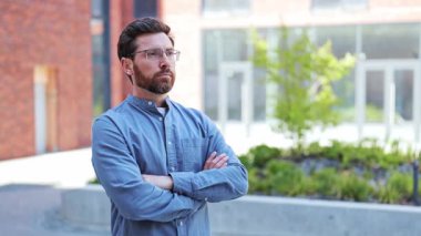 Gözlüklü ve günlük kıyafetleri olan kendine güvenen işadamı kollarını kavuşturmuş, derin düşüncelere dalmış bir şekilde ofis binasının yanında duruyor..