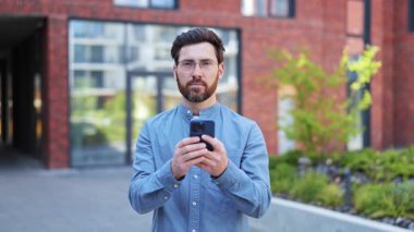 Şehir caddesinde kırmızı tuğlalı binanın yanında akıllı telefon kullanan gözlüklü ve sıradan giyinmiş yakışıklı işadamının portresi. Erkek mutlu, tatmin olmuş ve başarılı hissediyor..