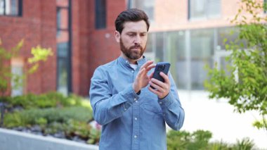 Sıradan kıyafetler içindeki yakışıklı iş adamı mutlu olur ve dışarıda başarıyı kutlar. Sakallı şık bir adam akıllı telefondan iyi haberlere sevinir..