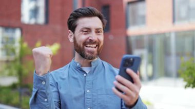 Başarılı şanslı iş adamı günlük kıyafetleriyle dışarıda yumruk sallayarak heyecanını ifade ediyor, başarıyı kutluyor. Neşeli bir adam telefonda iyi haberlerle mutlu ve hevesli..