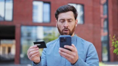 Günlük gömlekli şaşkın işadamı kredi kartı tutuyor ve telefona hayretle bakıyor. İş, finans ve teknoloji kavramı.