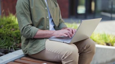 Yakışıklı, dışarıda bir dizüstü bilgisayarda çalışıyor. Parktaki bankta dizüstü bilgisayar kullanarak şık giyinen adam. Pozitif duygular.