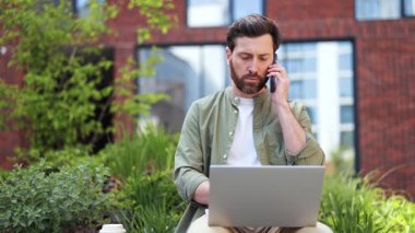 Sıradan giysiler içindeki odaklanmış iş adamı dizüstü bilgisayarla çalışıyor ve telefonla konuşuyor. Ofis binasının dışında oturan yakışıklı bir serbest yazar. Pozitif duygular, başarı.