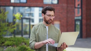 Dışarıda dizüstü bilgisayar kullanan, gözlüklü ve sıradan giyimli yakışıklı bir iş adamı. Arka plandaki modern ofis binası. İnsan mutlu olur..