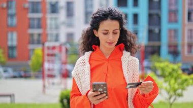 Turuncu tişörtlü neşeli bir kadın akıllı telefon ve online alışveriş yapan bir kredi kartı kullanıyor. Kolay ödeme işlemi.