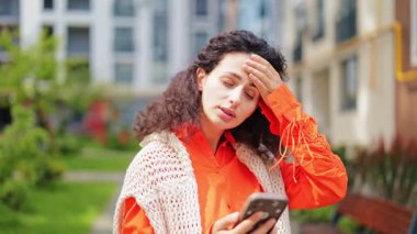 Turuncu gömlekli ve beyaz yelekli stresli kadın telefondaki kötü haberlerden rahatsız oluyor. Apartmanın dışında duruyor..