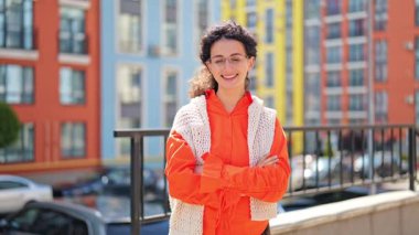 Parlak turuncu gömlekli, beyaz örgü yelekli neşeli genç bir kadın renkli evlerin yanında kollarını kavuşturarak kameraya gülümsüyor. Mutluluk ve neşe hissediyor..