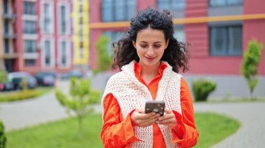 Şehirde telefon kullanan neşeli bir kadın. Turuncu gömlek ve beyaz örgü yelek giyen şık bir kadın internet iletişiminden hoşlanıyor.