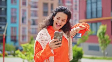 Turuncu tişört giyen şanslı kadın internet üzerinden kredi kartı ve telefon kullanarak ödeme yapıyor. Tarz sahibi bir kadın açık havada online mağazadan alışveriş yapıyor..