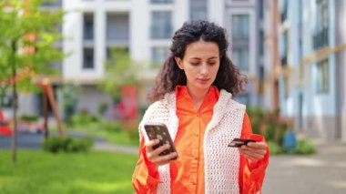 Kafası karışmış bir kadın akıllı telefondan online ödeme yapmada sorun yaşıyor. Turuncu tişörtlü şık bir kadın. Açık hava kredi kartı sorunu yüzünden hayal kırıklığına uğramış..