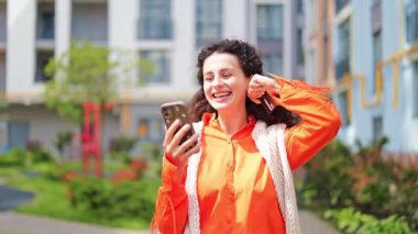 Turuncu tişörtlü ve beyaz şallı heyecanlı kadın telefon ve banka kartıyla online alışveriş yapıyor. Kendini mutlu hissediyor ve avluda alışveriş yapmaktan hoşlanıyor..