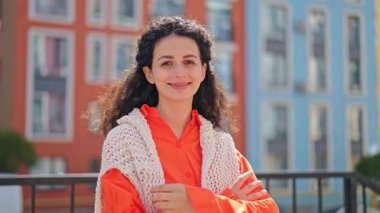 Şehir caddesinde kollarını kavuşturup gülümseyen turuncu elbiseli güzel, neşeli bir kadın. Mutlu, şık bir kadının portresi kendinden emin ve iyimser hissediyor..