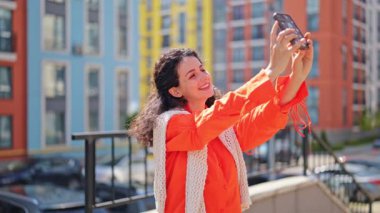 Neşeli esmer kadın şehirde akıllı telefonuyla selfie çekiyor. Şık ve rahat bir görünüm takınmıştı. Samimi bir gülümsemesi var ve mutluluğu ifade ediyor..