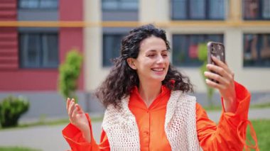 Turuncu elbiseli neşeli esmer kadın telefonunu kullanıyor, selfie çekiyor ve açık havada görüntülü sohbet ediyor..