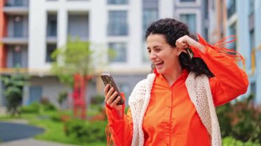Telefon ve kredi kartı kullanan turuncu tişörtlü ve beyaz şallı heyecanlı bir kadın. Kendini mutlu hissediyor ve dışarıda kutluyor. Kız bir şey almak için telefonunu ve kredi kartını kullanıyor..