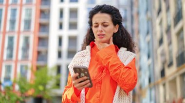 Ofis binasının yakınında akıllı telefon kullanan turuncu tişörtlü stresli kadın kötü haber alınca şaşırdı. Kıvırcık saçlı şık bir kadın açık havada endişeli..