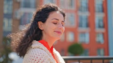 Gündelik giysiler içinde güzel bir kadın güneşli havada şehirde duruyor. Güneşin tadını çıkarıyor, mutlu ve huzurlu hissediyor..