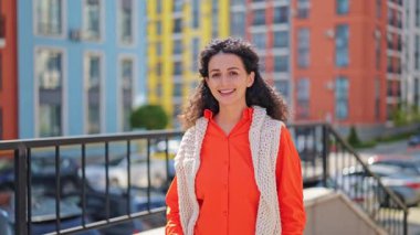 Parlak turuncu gömlekli neşeli kadın ve örülmüş yelek renkli binaların arka planına gülümsüyor..