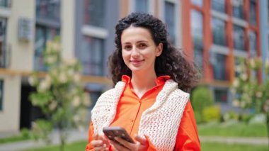 Turuncu gömlekli, beyaz örülü yelekli, elinde telefonla dışarıda gülümseyen neşeli genç bir kadın. Bulanık arka planda modern apartman ve yeşil çimenler..
