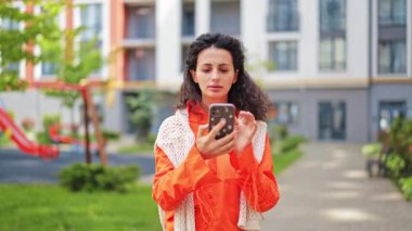 Telefon kullanan turuncu gömlekli neşeli bir kadın. Günlük giysiler içinde güzel bir bayan şehirde selfie çekiyor. Şık esmer mutlu ve rahat hissediyor..