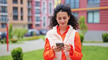 Akıllı telefon kullanan ve gülümseyen mutlu genç bir kız. Turuncu gömlekli, beyaz örgü yelekli şık bir kadın. Şehir parkı geçmişi.