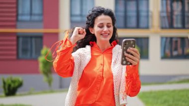 Dışarıda başarıyı kutlayan heyecanlı kadın. Turuncu ve beyaz elbiseli şık bir kadın, şehir ortamında telefon kullanarak neşe ifade ediyor. Olumlu duygular, başarı.