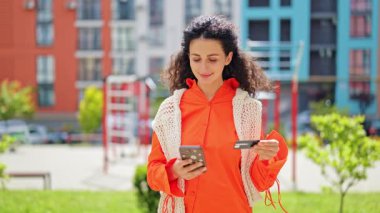 Akıllı telefon ve kredi kartıyla internetten alışveriş yapan neşeli bir kadın. Turuncu gömlekli şık bir bayan ve beyaz örgü bir yelek açık havada güvenli bir ödeme sağlıyor..