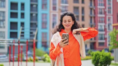 Heyecanlı kadın internette ödeme yapıyor. Şehirde akıllı telefon ve kredi kartı kullanan turuncu gömlekli neşeli bir kadın..