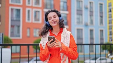 Turuncu elbiseli neşeli bir kadının portresi kulaklıkla müzik dinliyor ve şehir caddesinde telefon kullanıyor..
