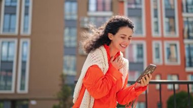 Modern bir binanın yanında duran heyecanlı kadın başarıyı kutluyor. Açık havada neşe ve mutluluğu ifade eder. Parlak günlük kıyafetler giyer..