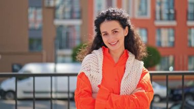 Şık turuncu gömlekli neşeli esmer kadın ve çapraz kollu örülü yelek gülüşü. Şehir caddesinde duran güzel bir kadının portresi. Olumlu duygular, neşe, mutluluk kavramı.