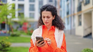 Hüsrana uğramış stil sahibi kadın modern binaların yakınında akıllı telefon kullanırken bir baş ağrısı hissediyor. Kıvırcık esmer saçları, turuncu gömleği ve beyaz örgü bir yeleği var..