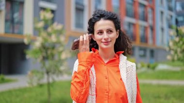 Havalı turuncu tişörtlü mutlu esmer kadın telefonda hoparlör kullanarak konuşuyor. Açık havada, modern binaların yanında ve geniş bir gülümseme içinde..