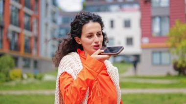 Akıllı telefon kullanan neşeli esmer kadın, dışarıya sesli mesaj yolluyor. Turuncu gömlekli, beyaz örülü yelek giyen şık bir kadın..