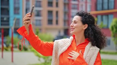 Turuncu gömlekli ve beyaz örgülü şallı neşeli kadın ev binalarının yanında selfie çekiyor ve mutlu bir şekilde gülümsüyor..