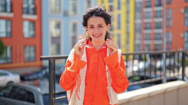 Şık turuncu gömlekli ve gözlüklü neşeli kadın şehirdeki renkli binaların yakınında poz veriyor. Gülümser ve hayattan zevk alır..