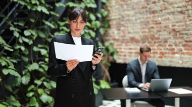 Kendine güveni tam bir iş kadını telefon ve belge okuyor. İş adamı arka planda dizüstü bilgisayarla çalışıyor. Duvarları ve bitkileri olan modern bir ofis..