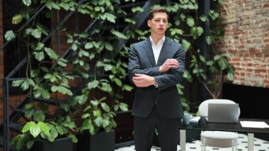 Takım elbiseli kendine güvenen işadamı modern ofiste kollarını kavuşturup yeşil bitkilerle ayakta duruyor. Başarılı ve gururlu hissediyor..