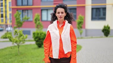 Parlak elbiseli, ciddi bir genç kadın, çapraz elli, bir iskan kompleksinin avlusunda duran jestleri durduruyor..