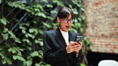 Heyecanlı takım elbiseli ve gözlüklü iş kadını telefonda iyi haberler alıyor. Başarılı kadın girişimci modern ofiste başarıyı kutluyor..