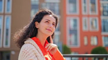 Turuncu gömlekli ve örgü şallı güzel bir kadın şehir binalarının arka planında duruyor, rüyasında yüzüne dokunuyor..